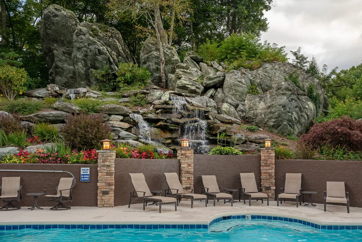 A stunning outdoor pool area with a natural rock formation, waterfall, and lounge chairs, surrounded by a landscaped garden. The setting is serene and inviting, perfect for swimming, sunbathing, or unwinding with friends.