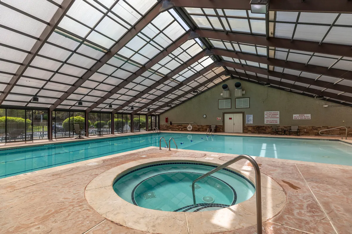 A serene indoor wellness area featuring a sparkling indoor pool, bubbling hot tub, glass ceiling that lets in natural light, and comfortable poolside lounge furniture. The atmosphere is tranquil and geared toward relaxation and rejuvenation.