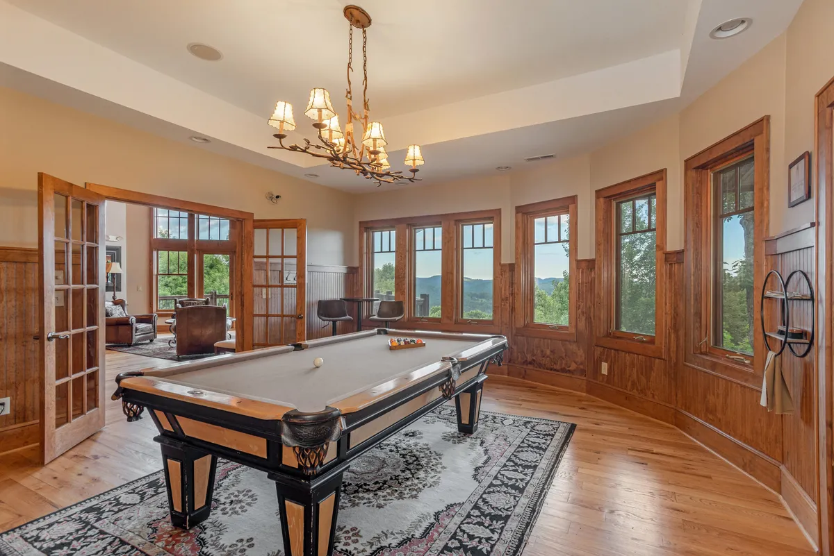 An interior scene of a cozy recreation area with a pool table, chandelier, large windows, and wood paneling, offering scenic mountain views. The space is designed for group activities and memorable shared moments.