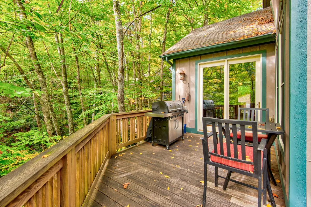 A tranquil deck scene with a gas grill, comfortable chairs, and a wooden railing looking out over leafy trees. The setup suggests easy access to outdoor cooking and socializing, with the grill ready for a barbecue and seating positioned for both intimate chats and group gatherings.