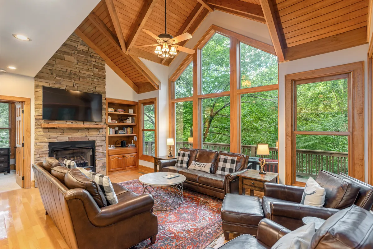 The image showcases a spacious, light-filled great room with soaring cathedral ceilings, rustic wooden beams, a striking stone fireplace, plush seating, and expansive windows that frame tranquil views of the surrounding forest. A decorative rug anchors the inviting seating area, creating a perfect space for relaxation or gathering with family and friends.