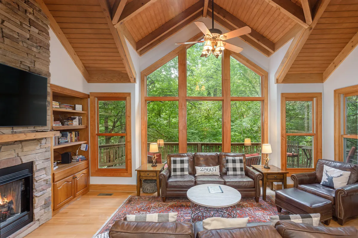 This image features a spacious great room with large windows overlooking the forest, comfortable seating, a stunning floor-to-ceiling stone fireplace, and a warm wooden ceiling. Natural light floods the space, and the arrangement clearly encourages family gatherings and relaxation, while offering tranquil nature views.