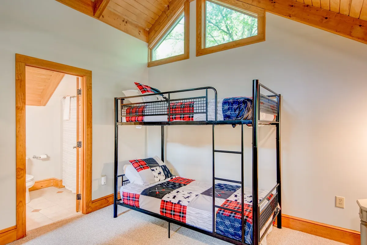 A cozy bunk room with a sturdy bunk bed, textured wooden ceiling, and large windows that let in ample light. The space is playful and inviting, capturing the spirit of a kid's paradise or a fun group retreat.