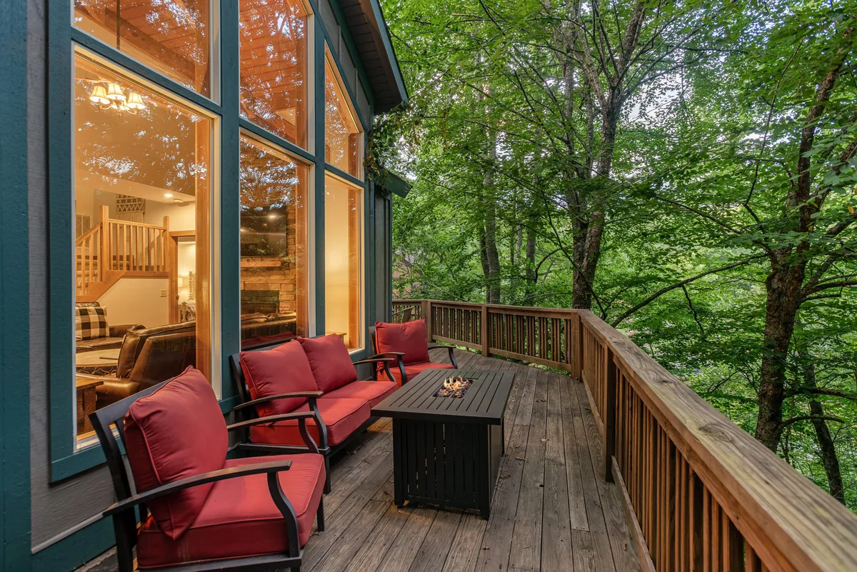 An exterior perspective showing the deck’s vibrant social scene with red cushioned chairs, a fire pit table, and large windows that connect indoor and outdoor spaces. The scene is lively yet welcoming, suggesting memorable evenings with friends and family while enjoying the fresh mountain air.