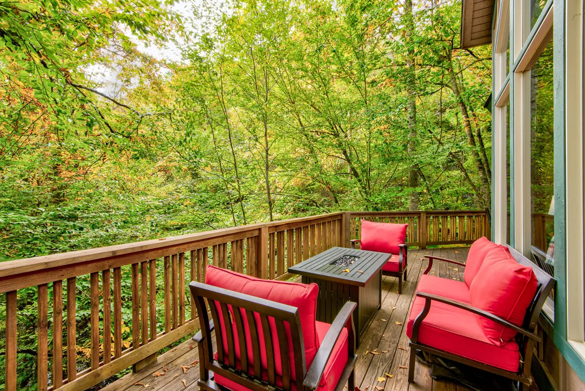 This image captures a wooden deck surrounded by tall trees, featuring red cushioned chairs arranged around a fire pit table. The deck radiates peacefulness, with soft evening light filtering through the trees, creating an inviting setting for relaxation or gatherings. The seating area and fire pit are the focal points, emphasizing comfort and sociability amidst nature.