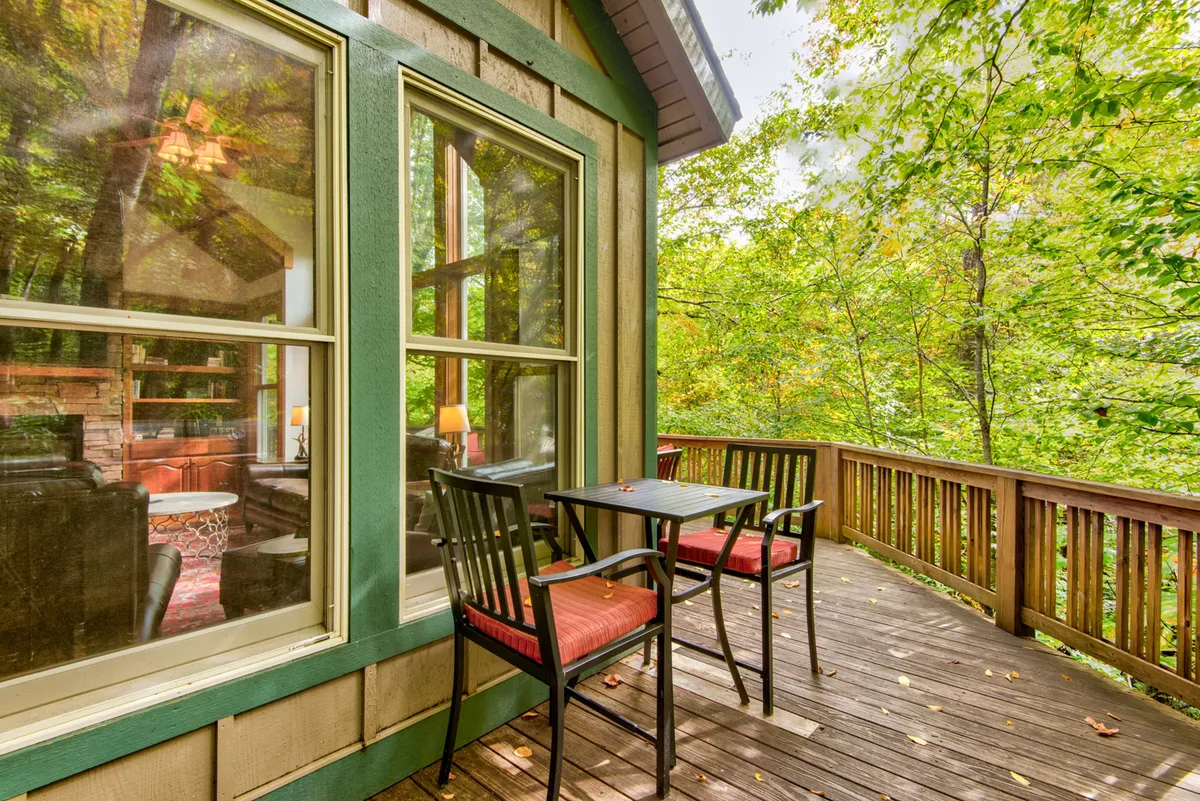 The image showcases a deck furnished with a table and chairs, large windows looking into the cozy interior, and vibrant greenery outdoors. The deck sits in a tranquil natural setting, ideal for relaxing after a day of adventure.