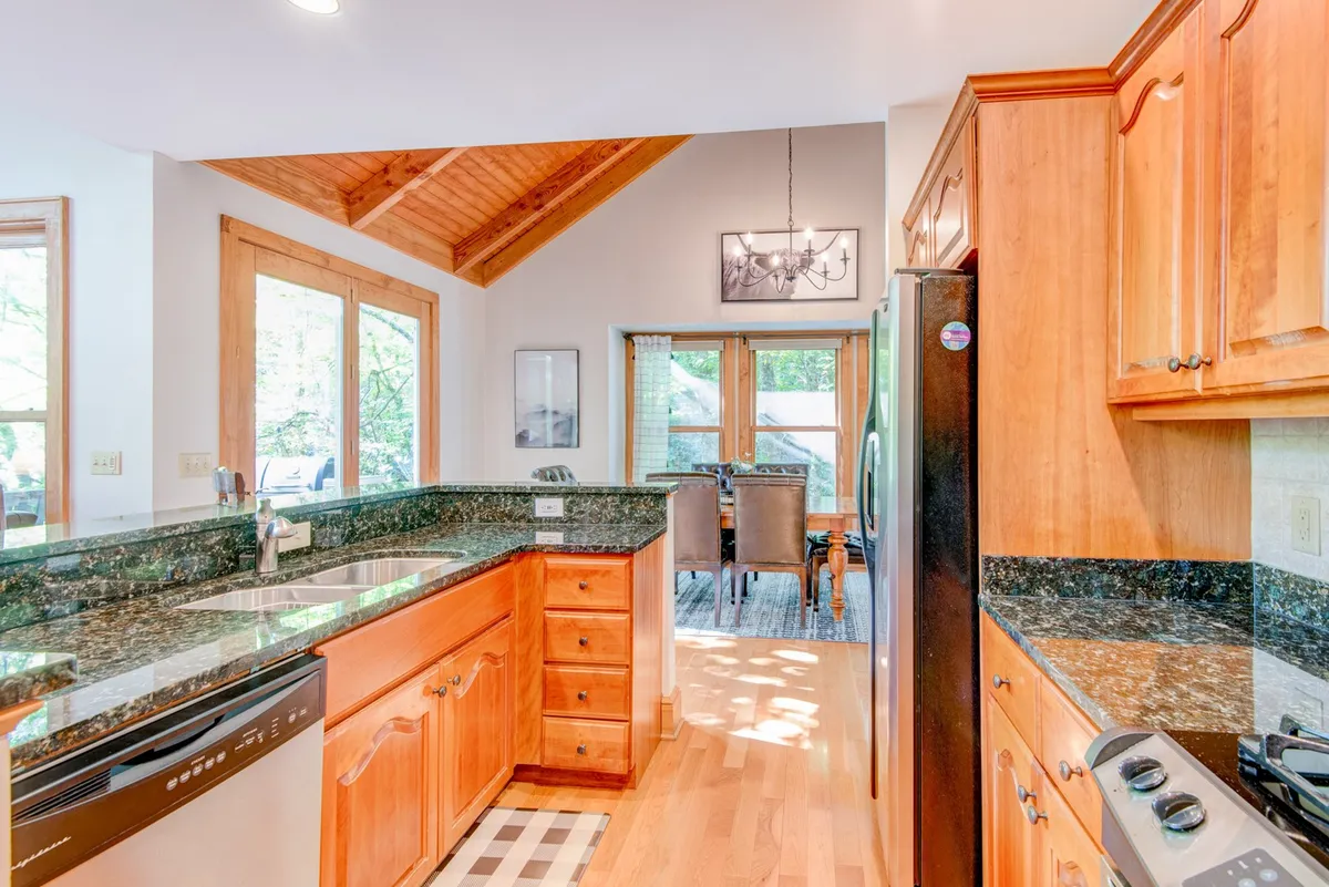 A warm, welcoming kitchen and dining area featuring granite countertops, wooden cabinetry, a large dining table, and generous windows that bring in mountain light. The layout seamlessly connects the kitchen to the dining and living spaces, perfect for entertaining and family meals.