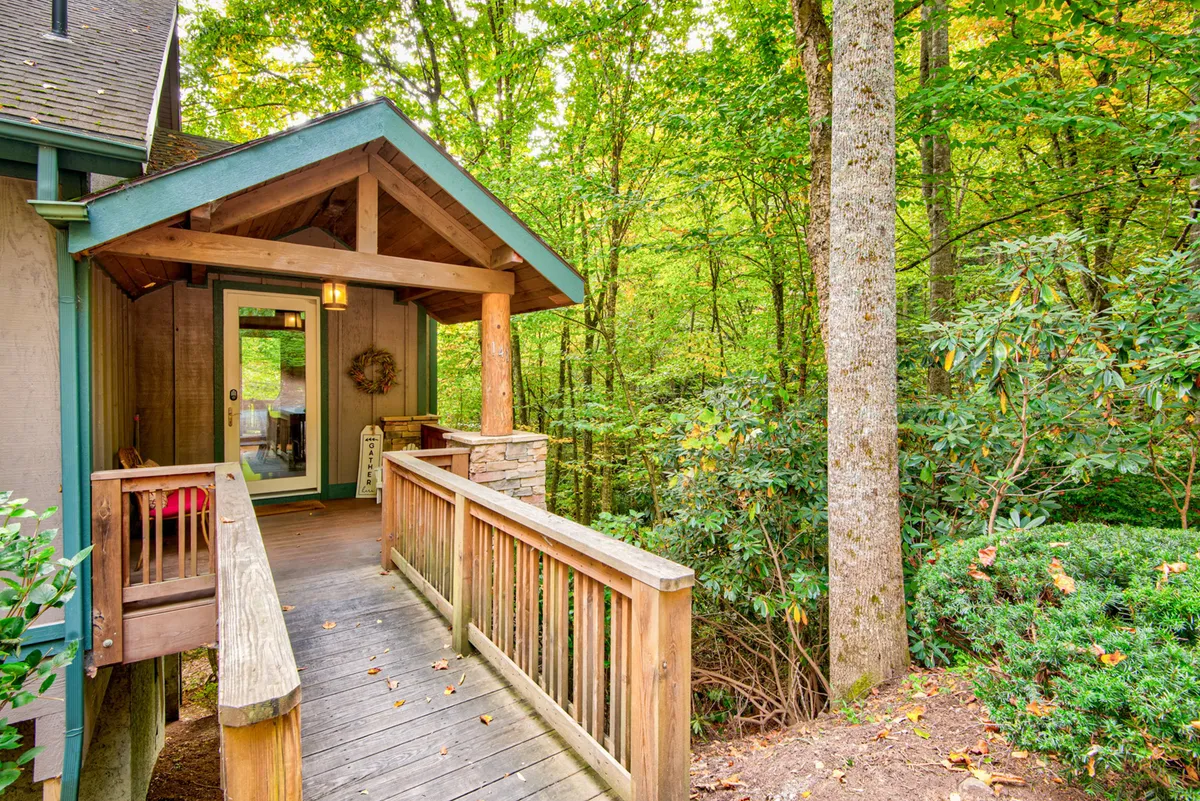 A welcoming exterior view featuring a wooden walkway, stone pillars, an entrance door with a decorative wreath, and greenery. The porch area is inviting and hints at a cozy, communal atmosphere for arrivals and departures.